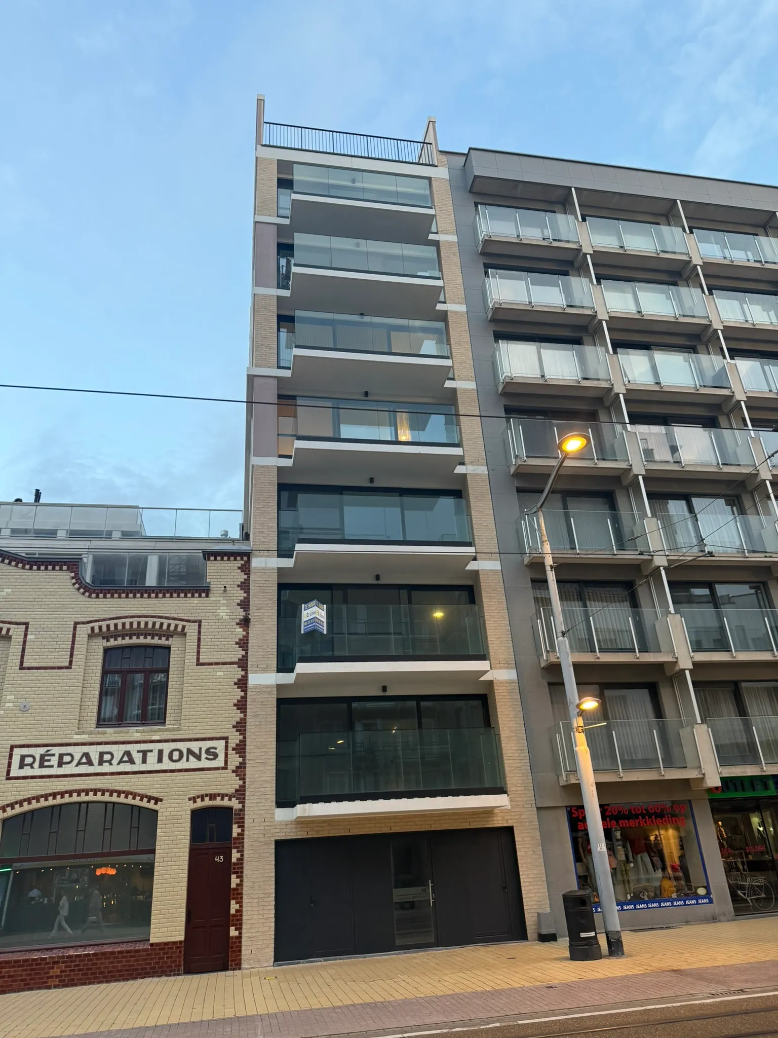 Afgewerkt appartementsgebouw van zeven bouwlagen met glazen balkons en lichte gevelsteen in een winkelstraat bij valavond