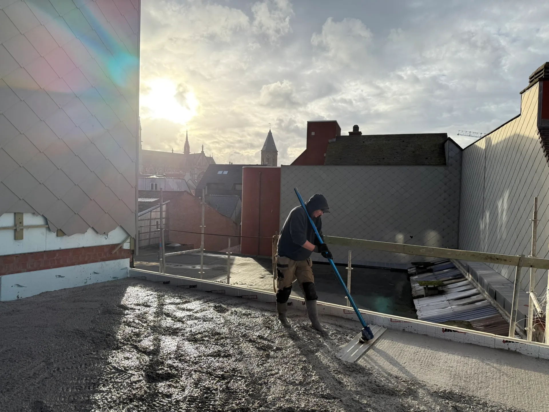 Vakman strijkt vers gestort beton af op een vloerplaat, met laagstaande zon en kerktorens op de achtergrond